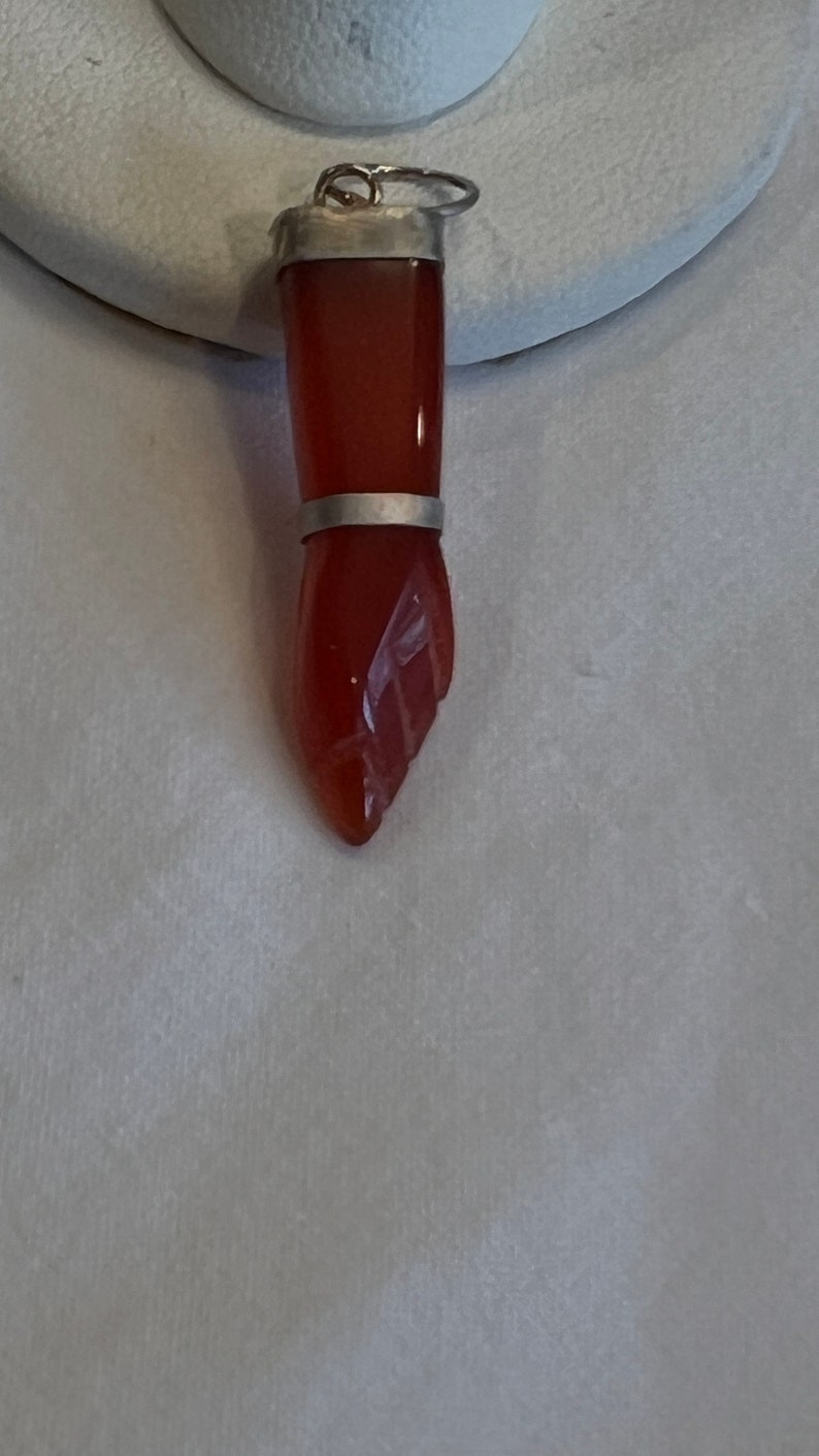 May include: A carved carnelian stone hand pendant with silver accents. The pendant is a detailed carving of a hand with a silver band around the wrist.