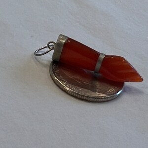 May include: A silver pendant with a carved carnelian stone in the shape of a hand. The stone is a reddish-orange color.