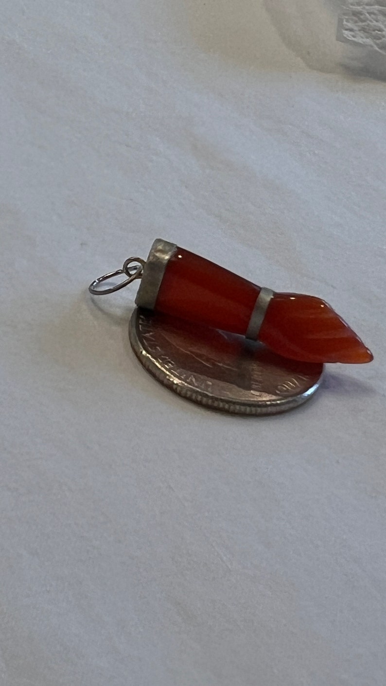 May include: A silver pendant with a carved carnelian stone in the shape of a hand. The stone is a reddish-orange color.