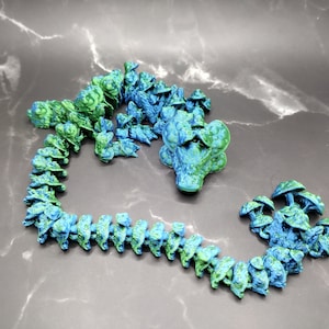 Könnte beinhalten: Eine blaue und grüne Plastikkette mit einem Drachenkopf-Anhänger. Die Kette besteht aus vielen kleinen, miteinander verbundenen Teilen, die wie Schuppen aussehen.