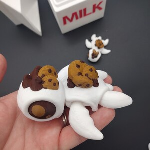Könnte beinhalten: Zwei weiße Keramikfiguren mit Schokoladen- und Keksdetails. Die Figuren sind in Form einer Schildkröte mit Flügeln gestaltet und halten Schokoladenkekse auf ihrem Rücken. Die Figuren sitzen auf einer Hand.