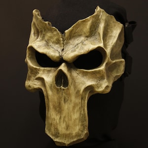 May include: A weathered, bone-coloured skull mask with large eye sockets and a prominent nose cavity. The mask has a textured surface and is worn against a black background. The mask is secured with a black strap.