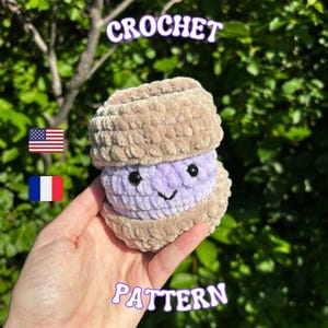 Peut inclure: Un petit jouet en crochet pelucheux et rond, composé d'un centre violet clair avec deux yeux en perles noires et un sourire brodé, pris entre deux couches texturées de couleur beige. Le texte CROCHET PATTERN est visible avec des drapeaux.