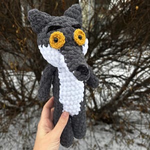 Peut inclure: Une poupée loup au crochet faite à la main, dans les tons de gris, de blanc et de noir. Le loup a de grands yeux jaunes et un long museau. La poupée est tenue devant un arrière-plan extérieur flou.