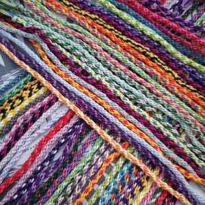 May include: A selection of brightly coloured friendship bracelets. The bracelets are made from braided threads in a variety of patterns and shades, including red, orange, yellow, green, blue, and purple. The image is a close-up of the bracelets.