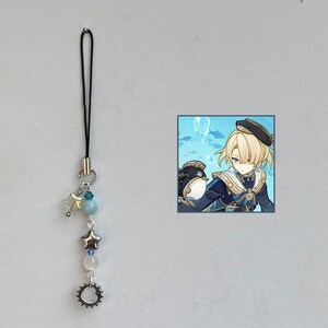 Könnte beinhalten: Ein silberner Charm mit schwarzem Kordel, mit einem Stern, Perlen und einem zahnradförmigen Charm. Ein kleines quadratisches Bild eines Charakters mit blonden Haaren und einem schwarzen Hut befindet sich neben dem Charm.
