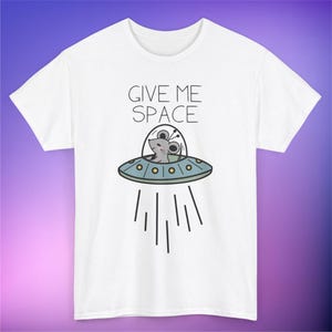 Könnte beinhalten: Weißes T-Shirt mit dem Text "GIVE ME SPACE" und einer Cartoon-Maus in einem Raumschiff. Die Maus ist grau, das Raumschiff hellblau mit gelben Akzenten. Schwarze Linien stellen die Bewegung des Raumschiffs dar.