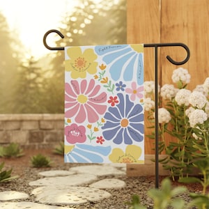 Può includere: Una bandiera da giardino colorata con sfondo bianco e un motivo floreale. I fiori sono rosa, blu, gialli e rossi. Il testo sulla bandiera recita "Fight Fearism, Justice & Courage, Be Relentless".