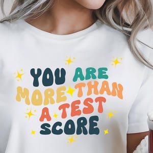 Könnte beinhalten: Weißes T-Shirt mit einer bunten Grafik im Retro-Stil, auf dem "You are more than a test score" steht.