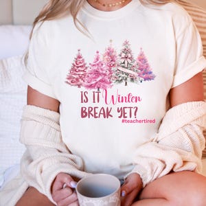 May include: Cream-coloured t-shirt with a design of pink Christmas trees. The text "IS IT Winter BREAK YET?" is printed below the trees, with the hashtag #teachertired. The shirt is made of a soft material.