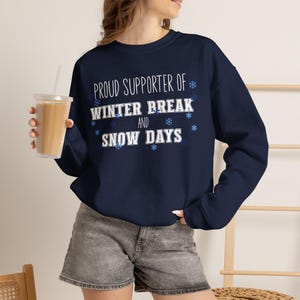 May include: A navy blue crewneck sweatshirt featuring the white text "PROUD SUPPORTER OF WINTER BREAK AND SNOW DAYS" with light blue snowflake graphics scattered around the lettering. The long-sleeved pullover has a ribbed collar and cuffs.