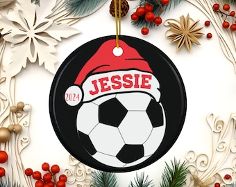 Op de afbeelding: Een zwart-witte voetbal met een rode kerstmanmuts op de bovenkant. De muts heeft de naam "JESSIE" erop geschreven. Het jaar "2024" staat aan de zijkant van de bal. Dit is een gepersonaliseerde kerstbal.