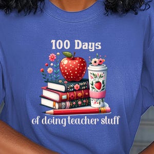 May include: A blue t-shirt with the text "100 Days of doing teacher stuff" in white. The design features a red apple with white polka dots, a stack of books with floral designs, and a coffee cup with a strawberry design.