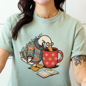 May include: A pale green t-shirt with a cartoon goose wearing glasses, a festive sweater, and a scarf, drinking from a red mug with snowflake patterns. The text "Is Christmas break yet?" is above the goose. An open book and pencil are at the bottom.