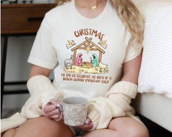 May include: Cream-colored t-shirt with a Christmas-themed graphic. The graphic depicts a nativity scene with the text "Christmas" above and "The day we celebrate the birth of a brown-skinned immigrant child" below. The shirt is short-sleeved.