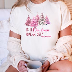 May include: Off-white t-shirt with a watercolour illustration of pink Christmas trees. The text "IS IT Christmas BREAK YET?" is printed in pink, with the hashtag #teachertired below. A comfortable, casual festive tee.