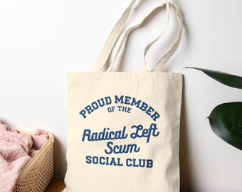 May include: Cream-colored tote bag with the text "PROUD MEMBER of the Radical Left Scum SOCIAL CLUB" in navy blue. The bag has two long handles and is made of a sturdy fabric. The bag is sitting on a wooden surface.