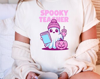 Puede incluir: Camiseta color crema con un diseño de un fantasma con gorro rosa, sosteniendo un libro y una bebida, con una calabaza rosa. El texto "SPOOKY TEACHER" está en rosa sobre el diseño.