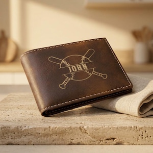 Könnte beinhalten: Ein braunes Lederportemonnaie mit Baseball-Design. Das Portemonnaie zeigt einen gravierten Baseball und Schläger mit dem Namen "John" in der Mitte. Das rechteckige Portemonnaie hat weiße Nähte an den Rändern.
