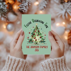Può includere: Un biglietto rettangolare verde chiaro con un'illustrazione centrale di un albero di Natale decorato con palline rosse e stelle. Il testo recita FESTIVE DECORATING TIME in alto e THE JOHNSON FAMILY 2026 in basso in rosso scuro.