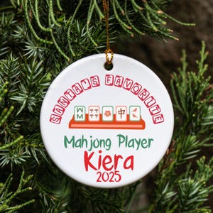 May include: A white ceramic Christmas ornament with the text "SANTA'S FAVORITE" in red. Below, it reads "Mahjong Player Kiera 2025". The ornament features Mahjong tiles and is hanging from a gold string.