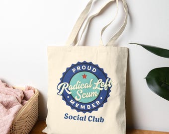 May include: Cream-colored tote bag with a blue and teal circular graphic that reads "Proud Radical Left Scum Member Social Club." The bag has two cream-colored handles and is made of a sturdy fabric. The bag is sitting on a wooden surface.