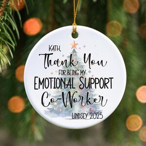 May include: A white ceramic Christmas ornament with a gold string for hanging. The ornament features the text "Kath, Thank You for being my Emotional Support Co-Worker" and "Lindsey 2025" with a watercolor Christmas tree design.