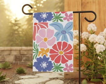 May include: A colorful garden flag with a white background and a pattern of pink, red, blue, yellow, and green flowers. The flag is attached to a black metal pole.
