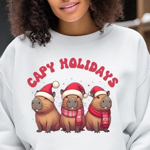 May include: White sweatshirt with a red graphic of three capybaras wearing Santa hats and the text "Capy Holidays".