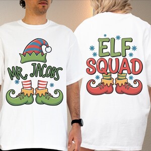 May include: White t-shirts with festive elf designs. One shirt has "Mr. Jacobs" with an elf hat and shoes. The other displays "Elf Squad" with elf shoes. Both designs incorporate blue snowflake accents.