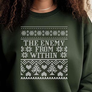 May include: Dark green sweatshirt with a white knit design. The design includes snowflakes, hearts, trees, and the text "THE ENEMY FROM WITHIN" in a bold font. The sweatshirt has a crew neck.