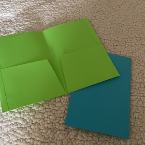 Puede incluir: Dos carpetas, una verde y otra azul, están sobre una superficie blanca y peluda. La carpeta verde está doblada por la mitad y tiene un bolsillo en la parte delantera. La carpeta azul está plana.