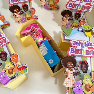 May include: A yellow number one shaped cardboard box filled with a pink Ring Pop and a blue snack package. The box is decorated with cutouts of a cartoon avocado, a purple character, and a girl, with text reading Jenyla's 1st Birthday.