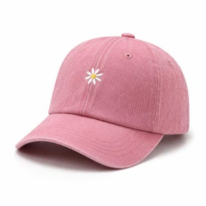 May include: A dusty rose baseball cap featuring a small, embroidered white daisy with a yellow centre on the front panel. The cap has a curved brim, stitched eyelets for ventilation, and a structured crown with a button at the top centre.