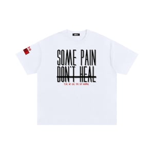 May include: White t-shirt with the text "SOME PAIN DON'T HEAL" in black, with a horizontal line through the text. Below the text is "YEAH, WE CALL THIS SH!T NORMAL" in red. A small red and white logo is on the left sleeve.