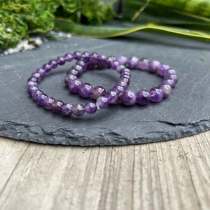 Pode incluir: Duas pulseiras de contas de ametista sobre uma superfície de ardósia cinza escura. As pulseiras são feitas de pequenas contas redondas roxas.