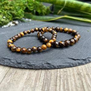 May include: Two brown and gold tiger's eye beaded bracelets on a black slate surface.