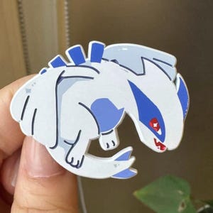 Può includere: Una spilla a cartone animato bianca e blu di una creatura mitica con occhi rossi e un becco affilato. La spilla è tenuta da una mano su uno sfondo sfocato. La creatura ha un design stilizzato.