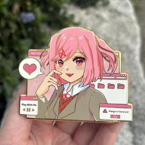Puede incluir: Un pin de personaje de anime de pelo rosa con borde dorado. El pin presenta un personaje con cabello rosa, ojos rojos y uniforme escolar. El texto en el pin incluye "Play With Me" y "Manga is literature!" con iconos de corazón y carpeta.