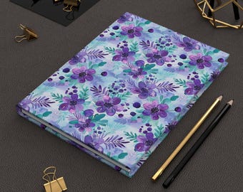 May include: A purple and blue floral patterned notebook with a white background. The notebook is closed and lying on a gray surface.