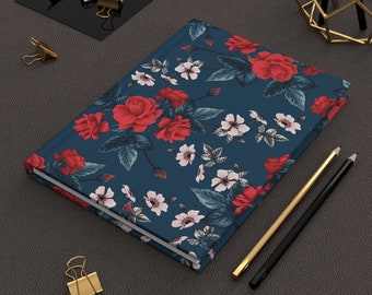 May include: A hardcover journal with a navy blue cover adorned with a floral pattern of red roses and white blossoms. The journal is accompanied by gold and black pencils and gold binder clips.
