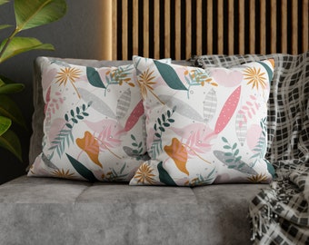 May include: Two square throw pillows with a white background and a colorful botanical pattern. The pattern includes pink, orange, teal, and gray leaves and floral elements. The pillows are resting on a gray sofa.