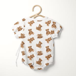 May include: A white short-sleeved bodysuit featuring a repeating pattern of brown teddy bears. The garment is made of a textured fabric and includes side tie closures. It is displayed on a light-colored wooden hanger against a plain background.