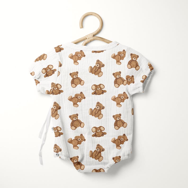 May include: A white short-sleeved bodysuit featuring a repeating pattern of brown teddy bears. The garment is made of a textured fabric and includes side tie closures. It is displayed on a light-colored wooden hanger against a plain background.