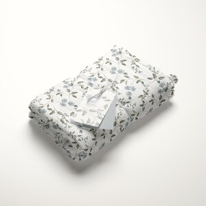 May include: A white fabric gift wrap with a delicate floral pattern featuring blue flowers and green leaves. The fabric is folded and tied with a white tag.