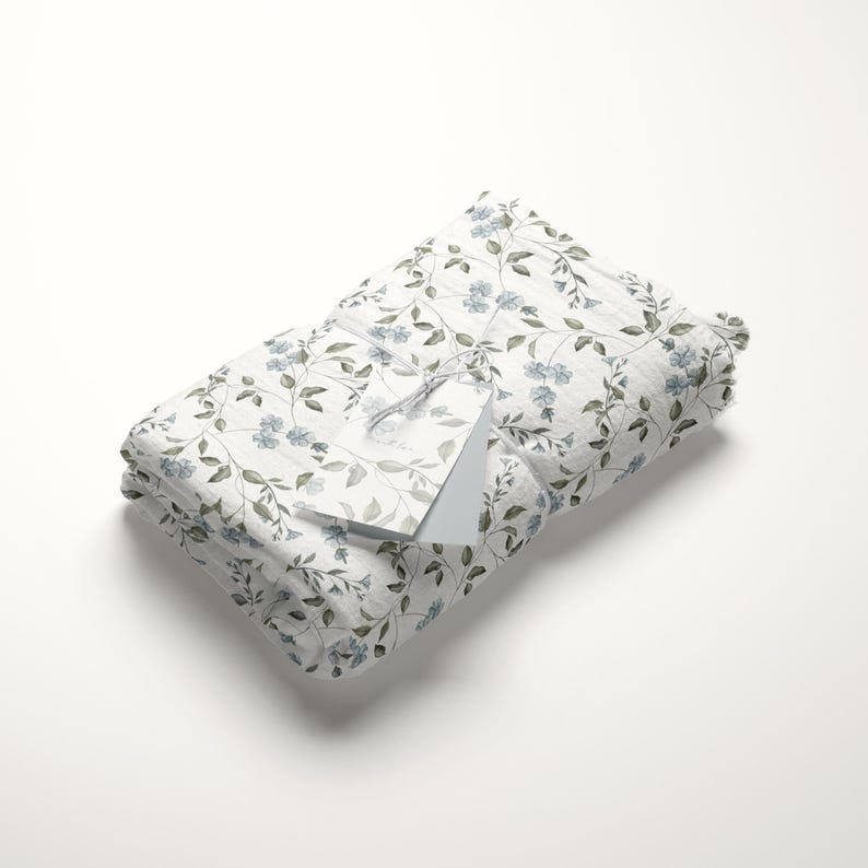 May include: A white fabric gift wrap with a delicate floral pattern featuring blue flowers and green leaves. The fabric is folded and tied with a white tag.