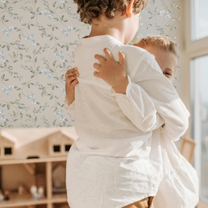 May include: Two children, one wearing a white shirt, hug each other in front of a light brown floral wallpaper. The child in the front is holding the other child's arm.