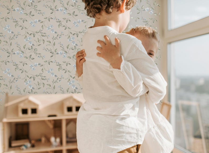 May include: Two children, one wearing a white shirt, hug each other in front of a light brown floral wallpaper. The child in the front is holding the other child's arm.