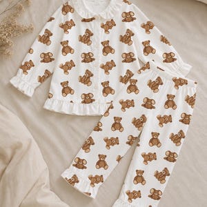 May include: A white long-sleeve pajama set featuring an all-over brown teddy bear print. The top has a button-down front, a rounded collar with ruffles, and ruffled sleeve cuffs. The matching pants feature an elastic waistband and ruffled hems.