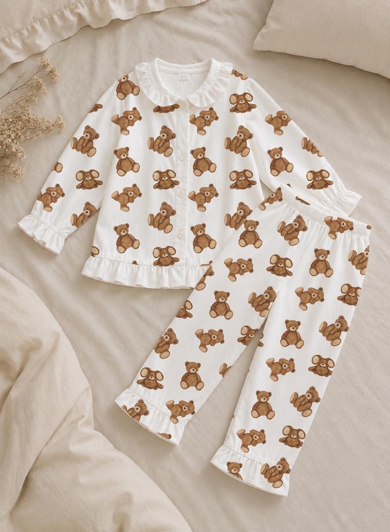 May include: A white long-sleeve pajama set featuring an all-over brown teddy bear print. The top has a button-down front, a rounded collar with ruffles, and ruffled sleeve cuffs. The matching pants feature an elastic waistband and ruffled hems.